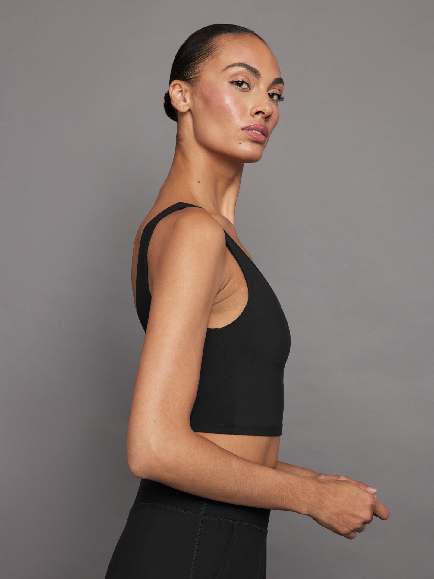 Model wears a black, longline sports bra with a deep V-neck front and an open V-shaped back, made from sleek, smooth fabric.
