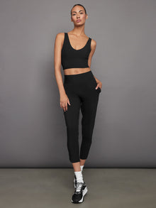 Model wears a black, longline sports bra with a deep V-neck front and an open V-shaped back, made from sleek, smooth fabric.
