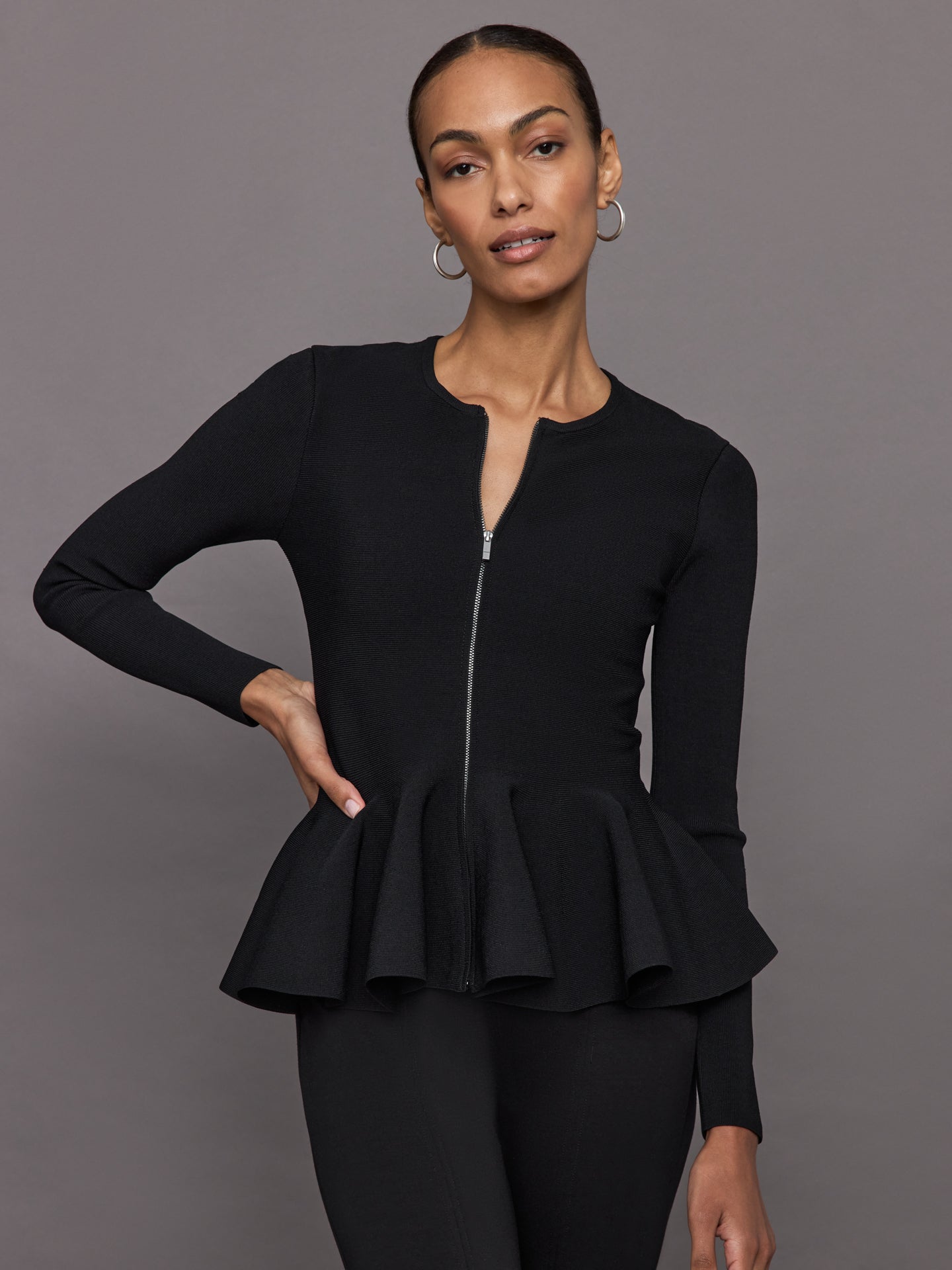 Model wears black long-sleeve zip-up sweater with a fitted bodice and flared peplum hem.