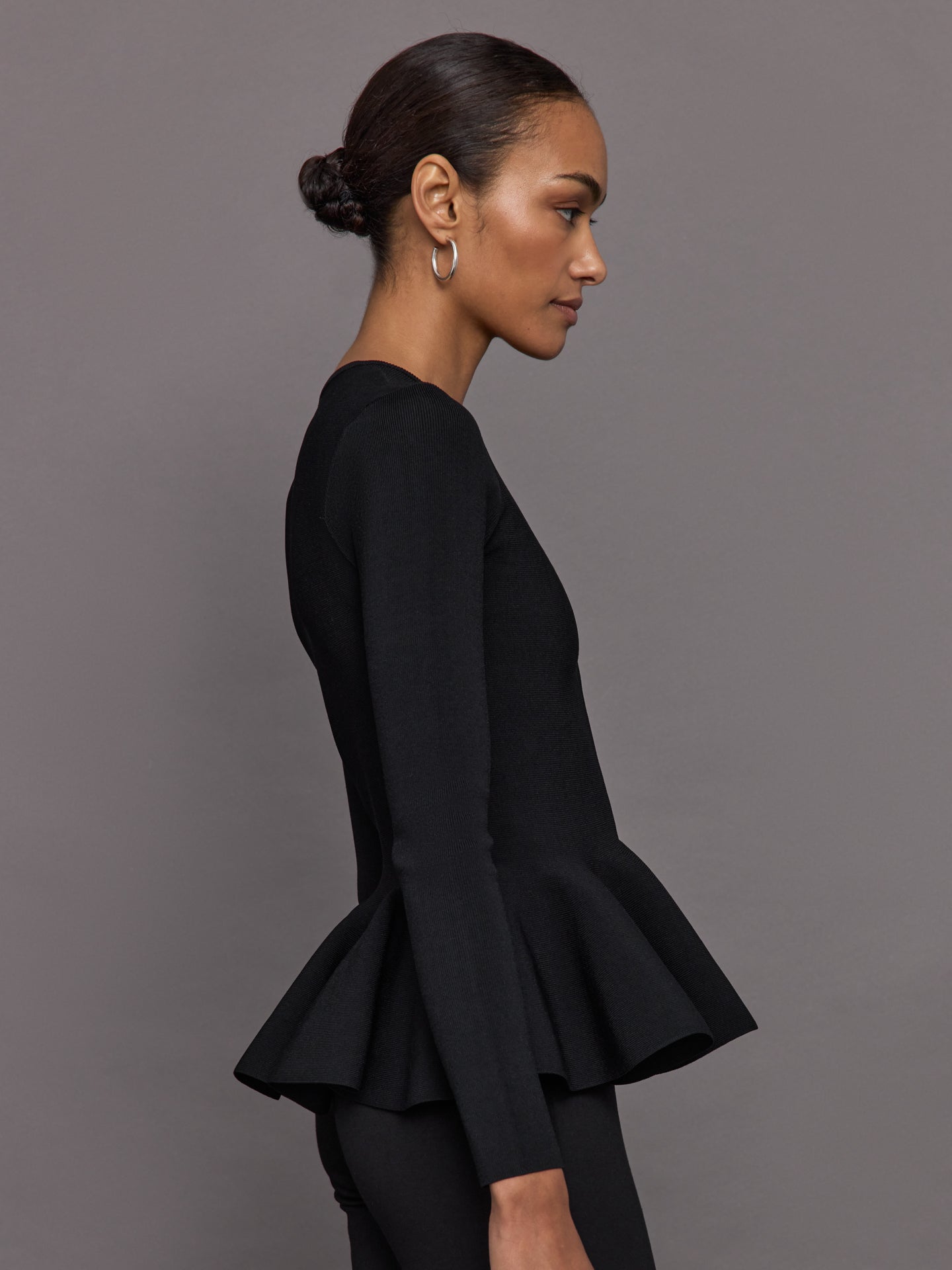 Model wears black long-sleeve zip-up sweater with a fitted bodice and flared peplum hem.