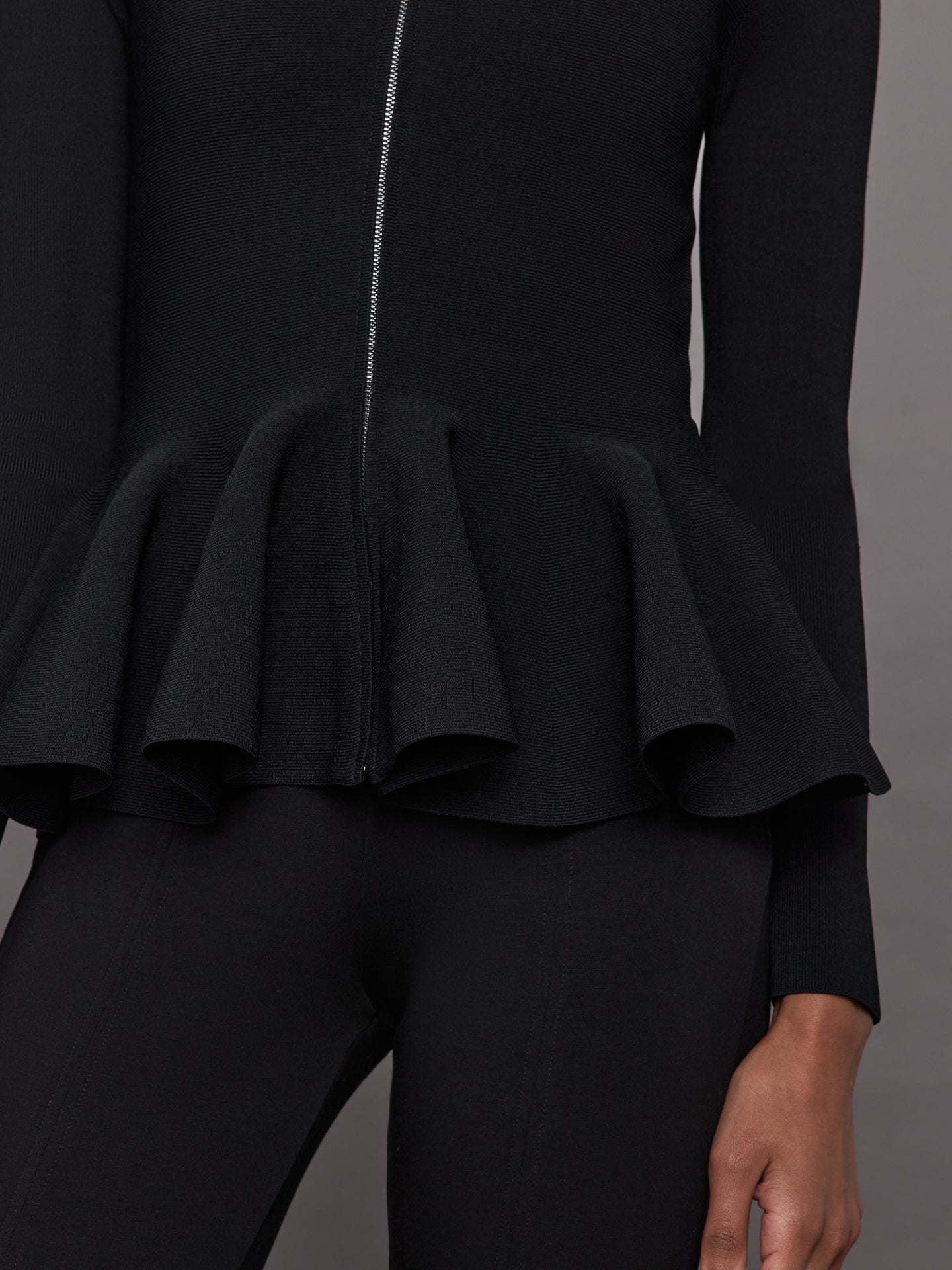 Model wears black long-sleeve zip-up sweater with a fitted bodice and flared peplum hem.
