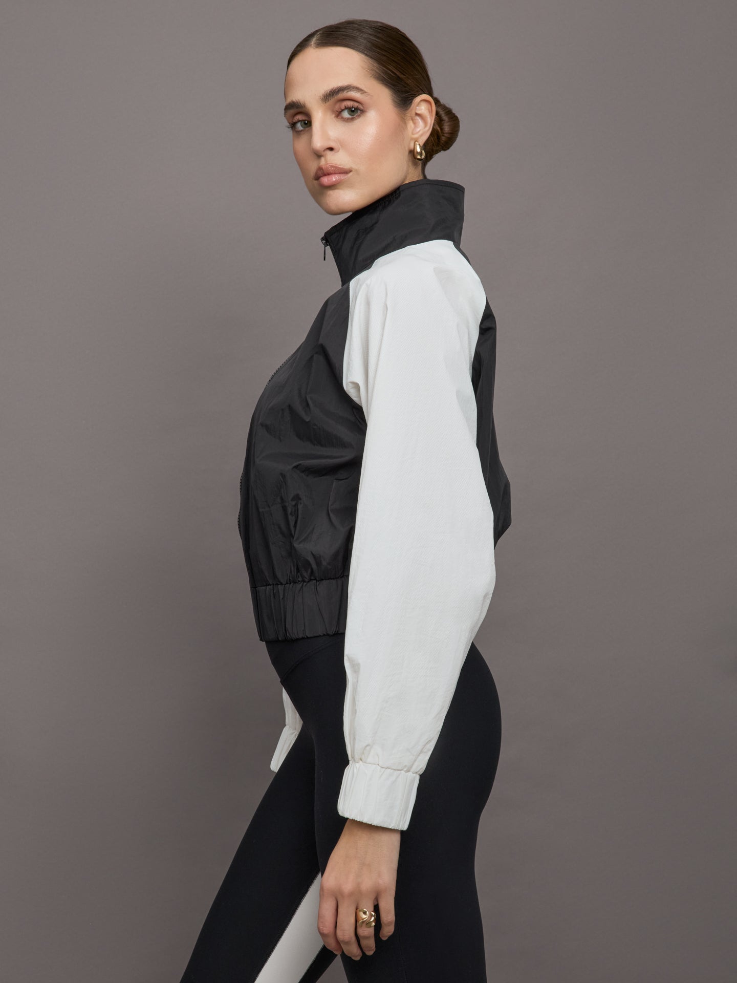 Model wears a cropped windbreaker with a popped collar, black bodice and white long sleeves with a cinched wristline.
