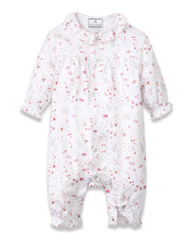 The Baby's Twill Madeline Romper in Love Song is a white long-sleeve romper with ruffled cuffs and collar, decorated with a pink pattern of hearts, glasses, and small animals. It features snap buttons down the front and legs.