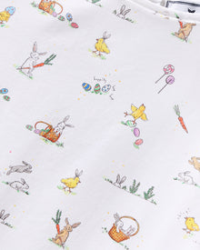 A young girl with short brown hair smiles in the Girl's Twill Amelie Nightgown in Easter Frolic, a white sleeveless dress featuring playful animal and nature prints, standing against a plain light background.