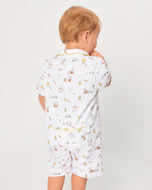 A young child with blonde hair wears the Baby's Twill Summer Romper in Easter Frolic—white with colorful animal prints and yellow trim—standing against a plain light background, holding two wooden blocks and looking to the side.