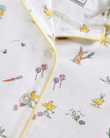 A young child with blonde hair wears the Baby's Twill Summer Romper in Easter Frolic—white with colorful animal prints and yellow trim—standing against a plain light background, holding two wooden blocks and looking to the side.