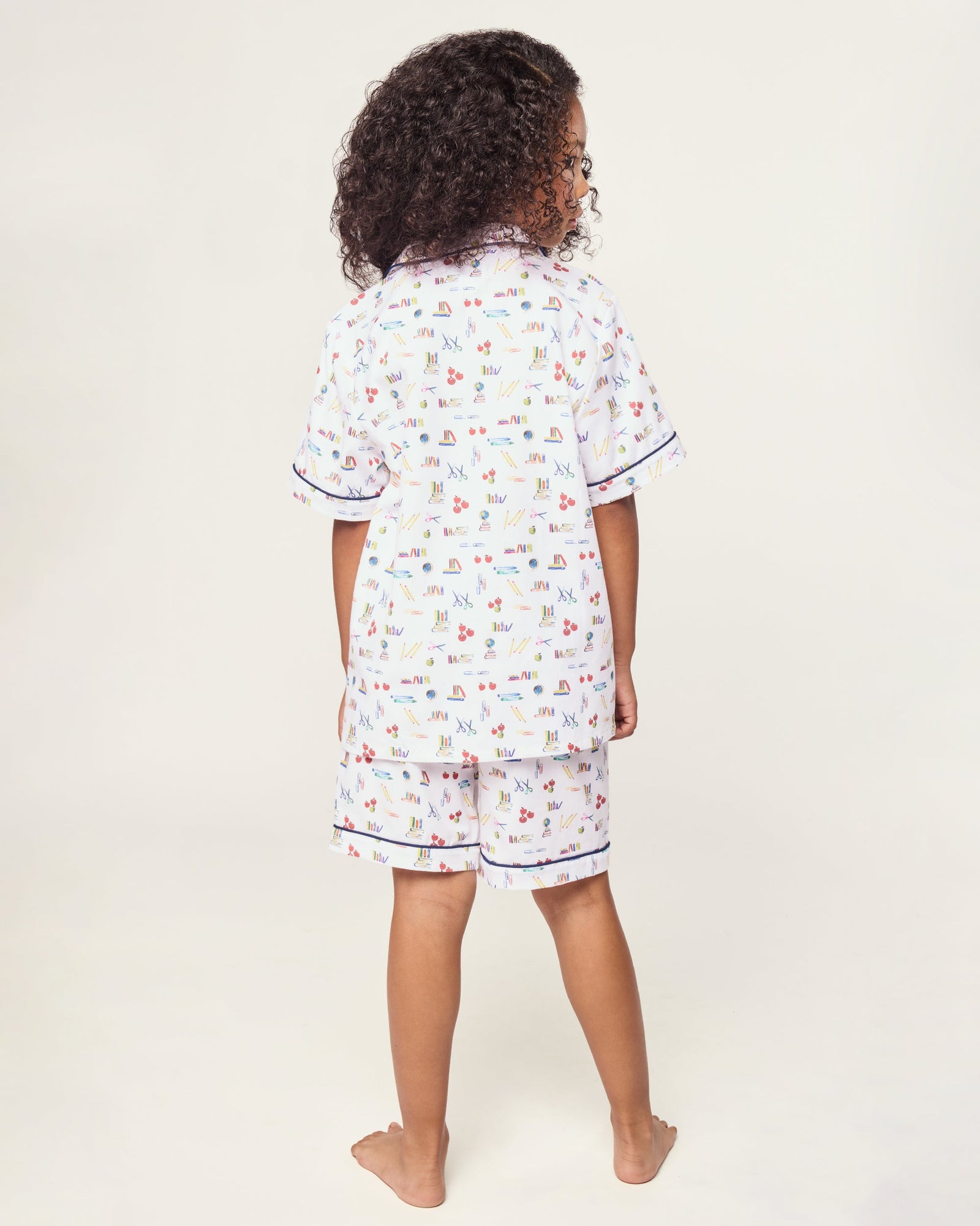 The Kid's Twill Short Set in Literary Lessons features a white base with colorful school-themed prints—pencils, books, and erasers—plus navy piping on the collar, pocket, sleeves, and shorts.