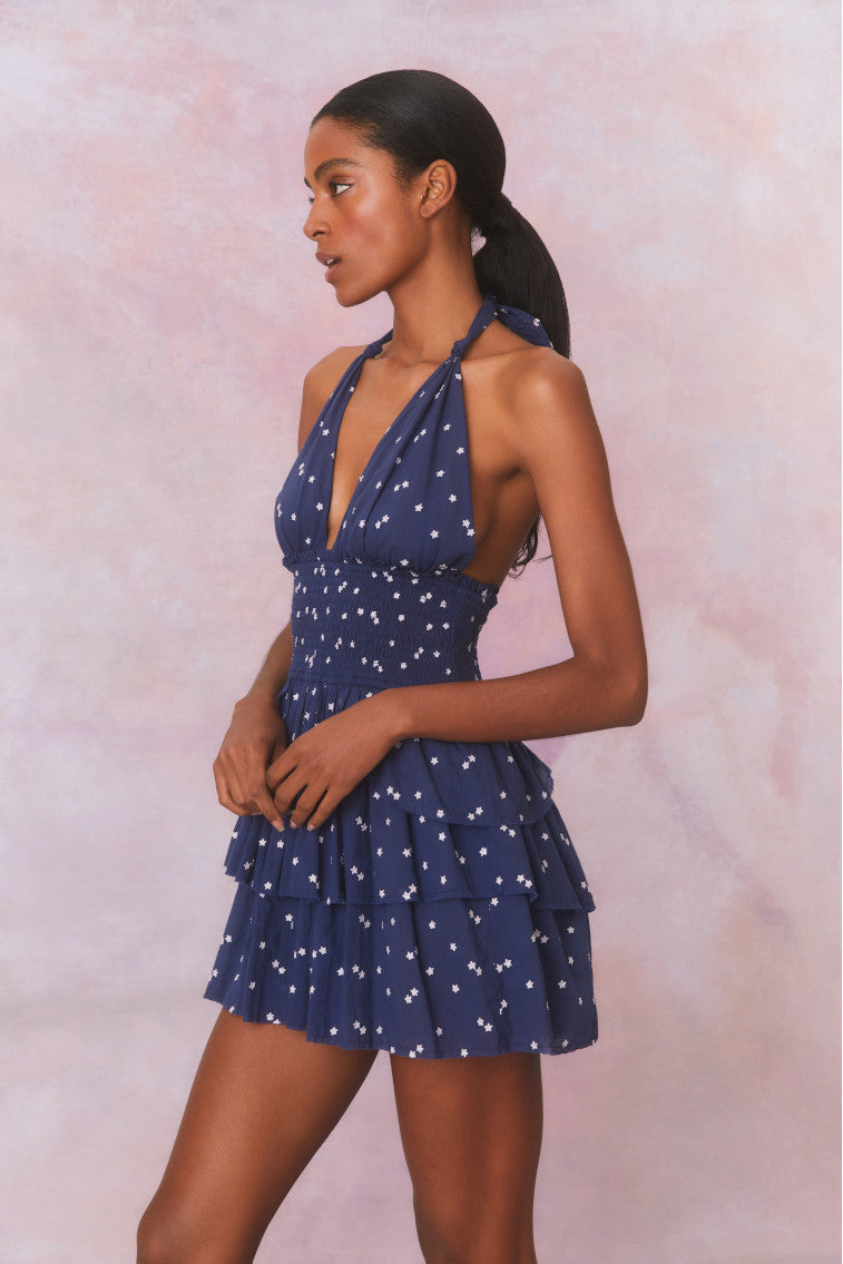 Low-cut navy blue convertible halter dress with hand-painted star print all over and a smocked waist, ties in a generous bow at the neck and can be crossed at front with a flared skirt with two ruffled tiers.