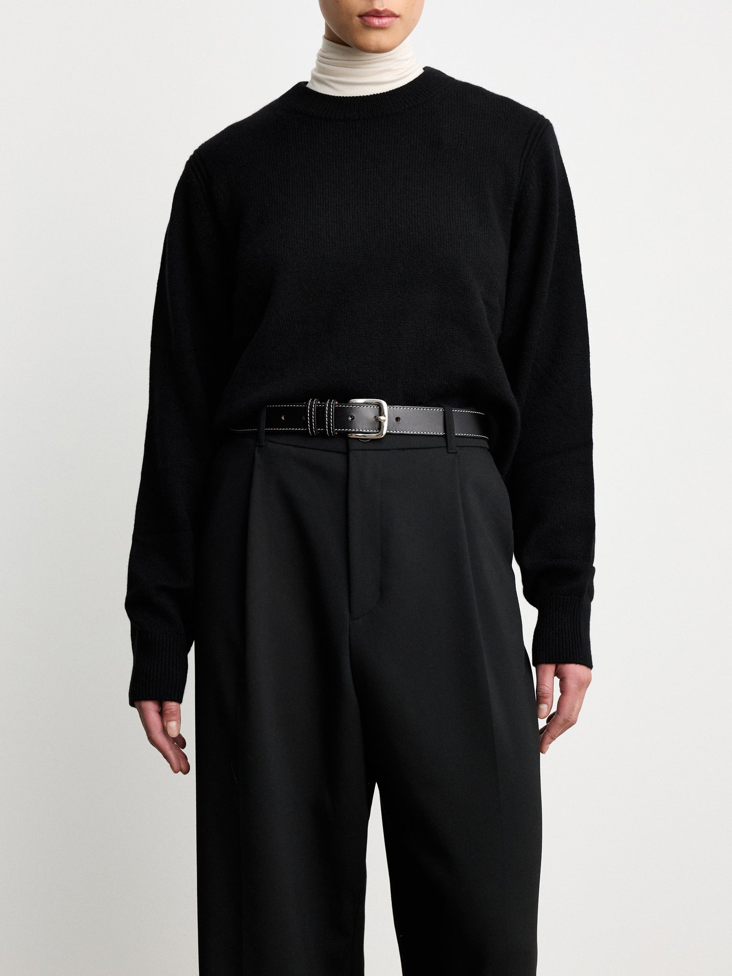 Model wearing D√©hanche Louison black leather belt with white stitching and silver buckle, styled with black trousers and a black sweater