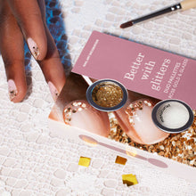 A hand with nude nails, adorned with Manucurist’s Glitters Duo Rose Gold  Glazed, rests on lace beside a pink and white 