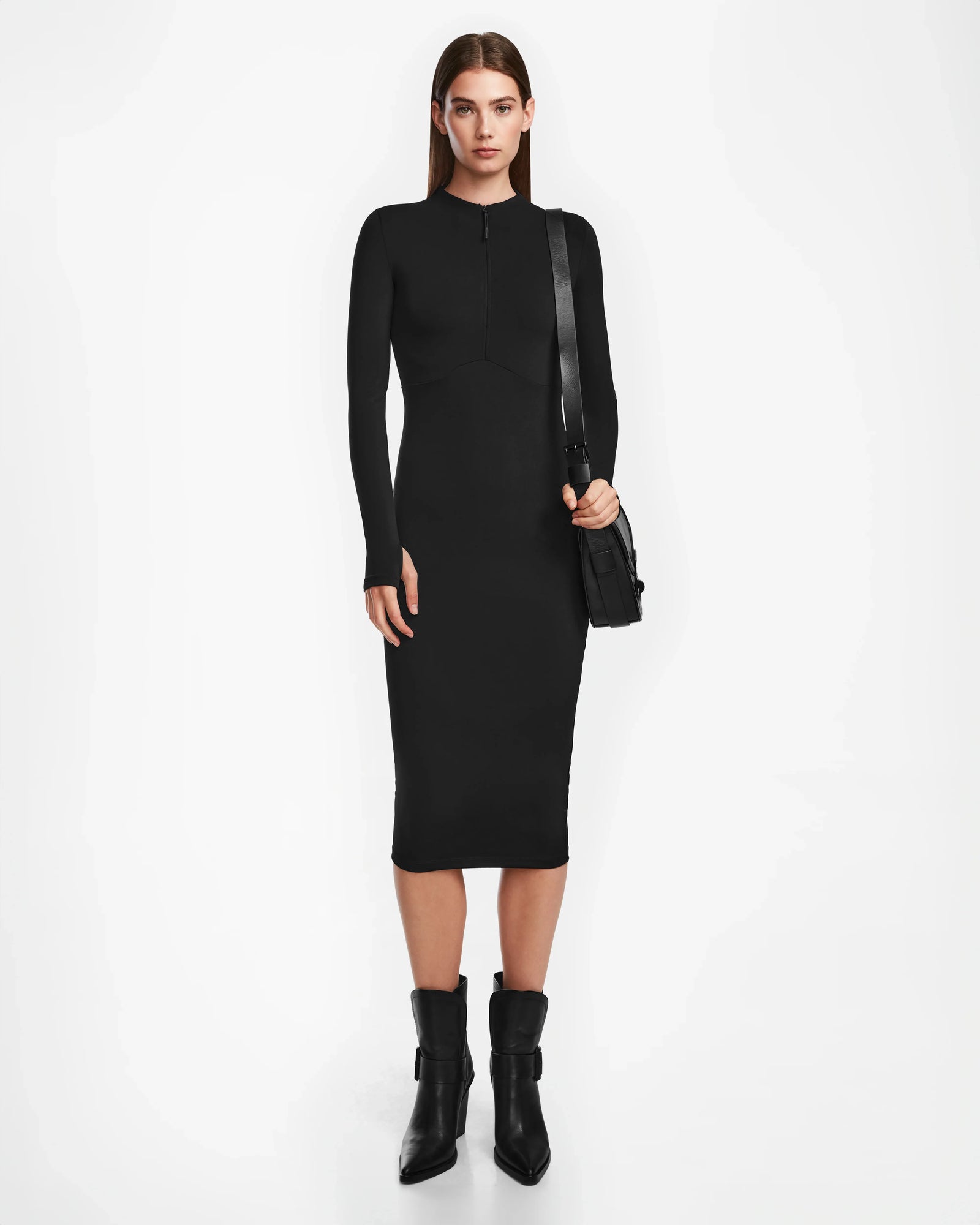 A woman faces forward wearing the Rudsak AGNES - 8524551 fitted long-sleeve black stretch dress, paired with black ankle boots and a black shoulder bag. She has straight brown hair and poses against a plain white background.