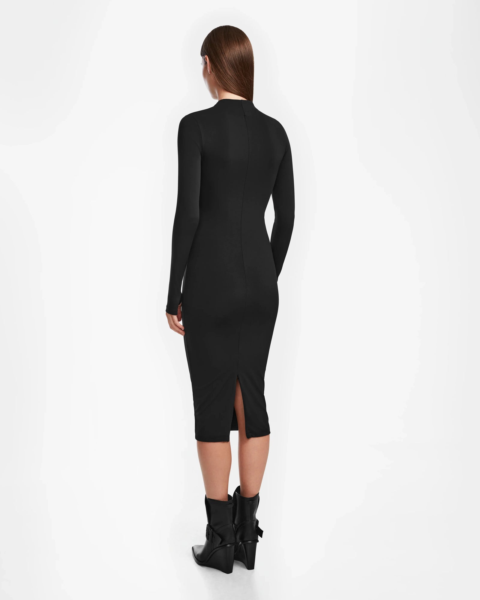 A woman with straight brown hair stands facing away, wearing the Rudsak AGNES - 8524551 Black, a fitted long-sleeve black womens bodycon stretch dress with a back slit, paired with black ankle boots against a plain white background.