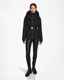 A woman faces forward wearing the Rudsak ALISA - L124506 Black quilted down puffer with vegan leather belt, black shiny leggings, and black boots. The RDS-certified jacket is showcased against a plain white background.