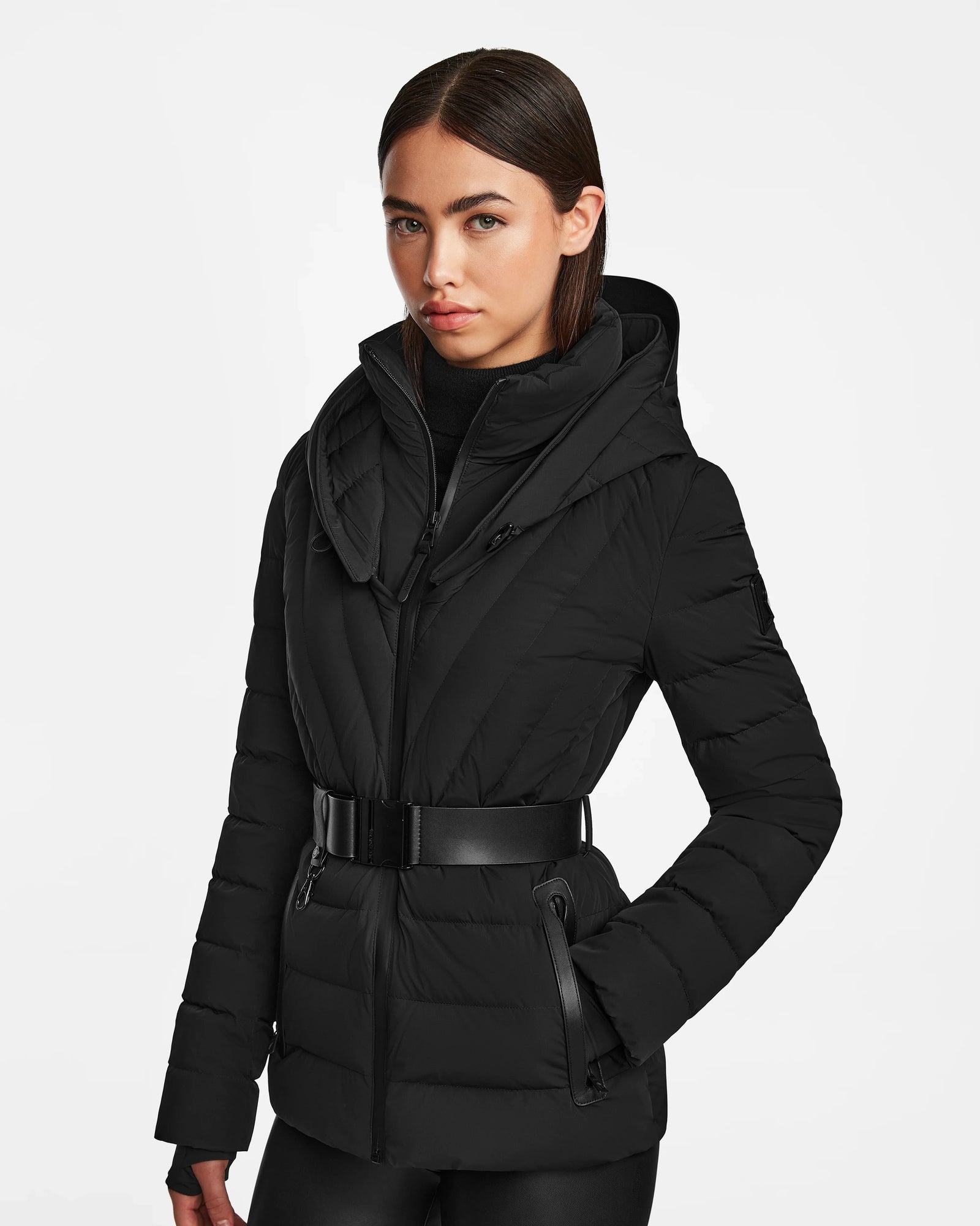 A woman with straight dark hair wears the Rudsak ALISA - L124506 Black, a womens quilted down puffer with vegan leather belt, high collar, and hood. She stands against a white background with hands in pockets and a neutral expression.