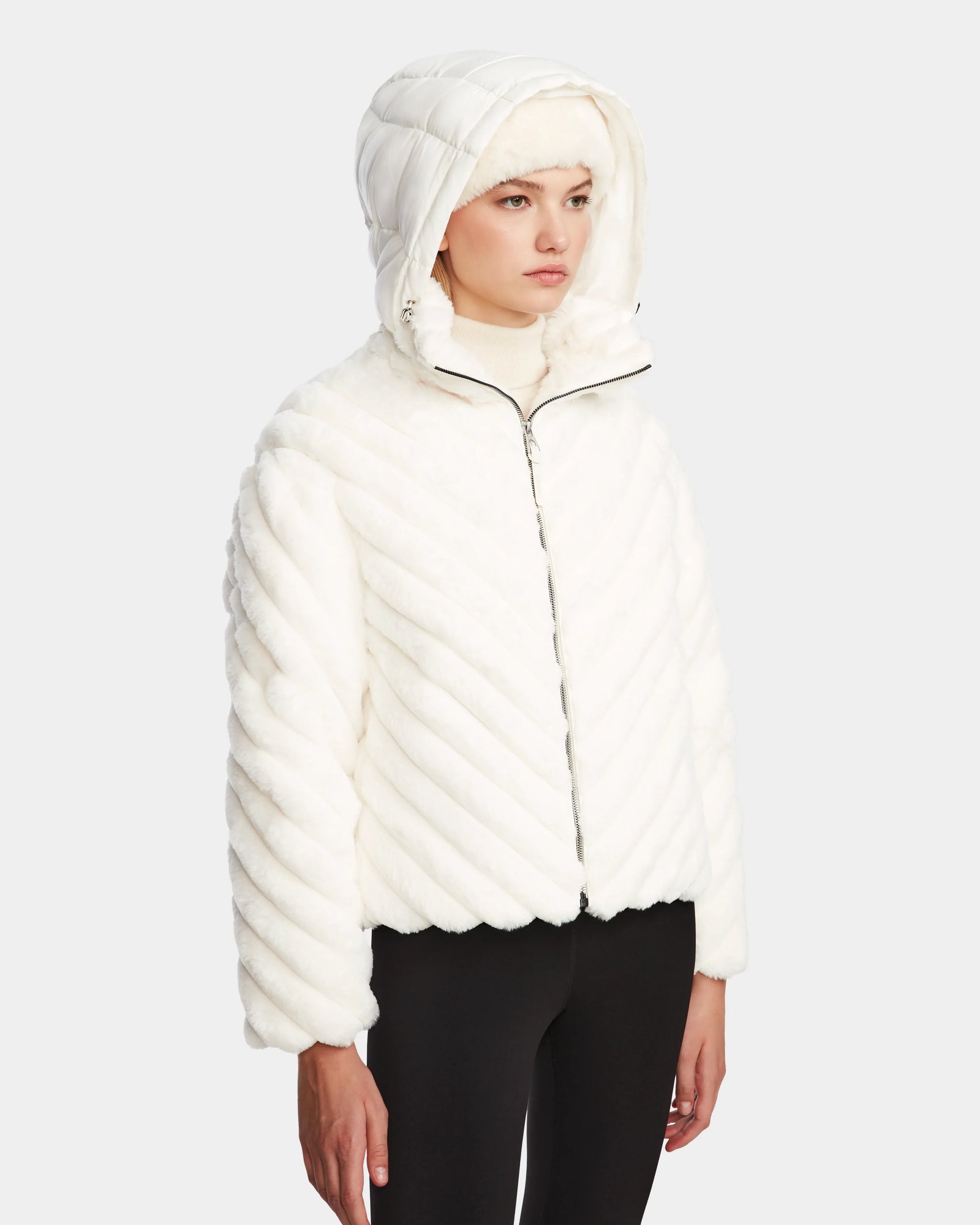 Someone wearing the Rudsak ANASTASIA - 8124804 Iceland white, hooded, RDS-certified down quilted jacket with diagonal stitching and black pants stands against a plain light gray background.