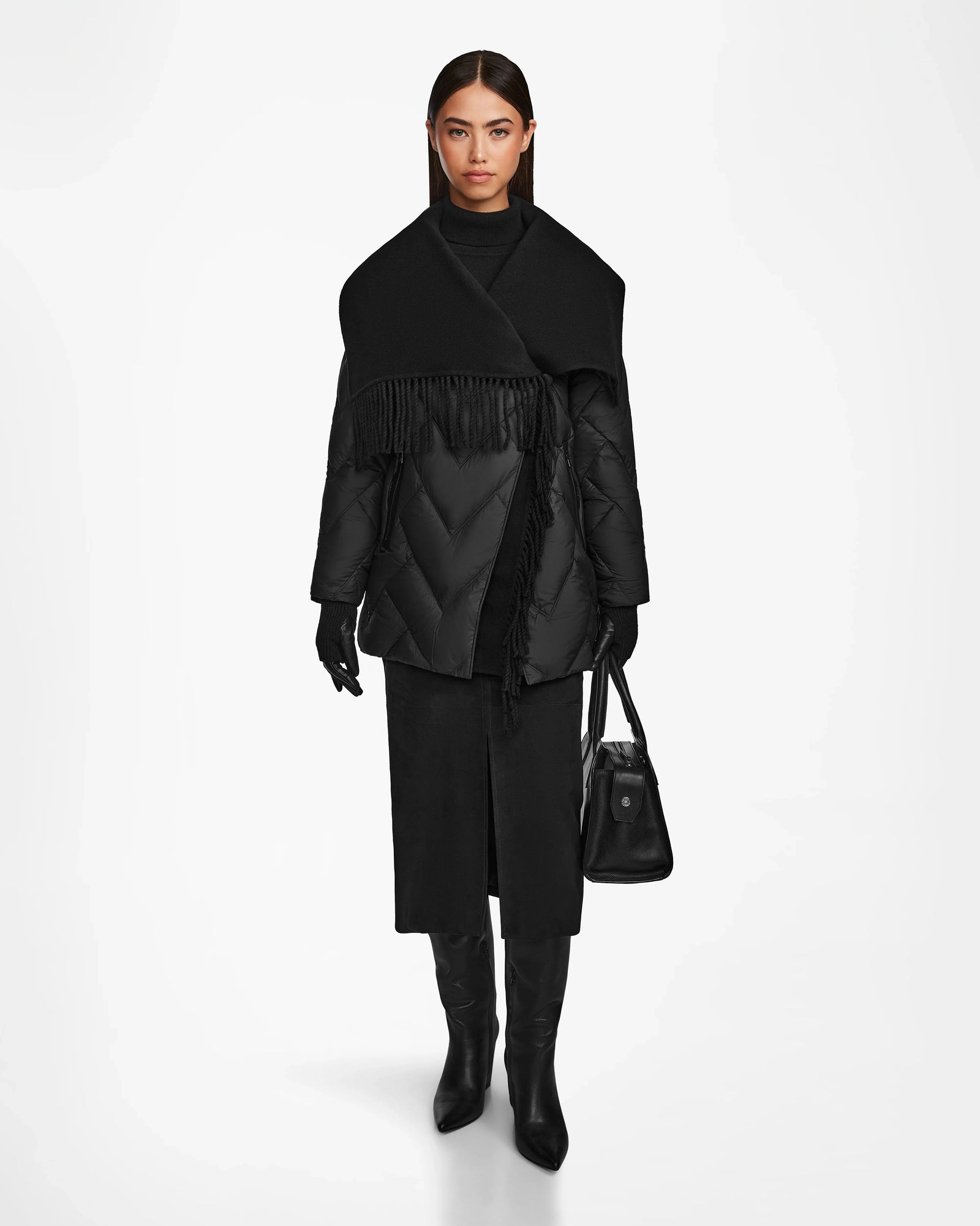 A woman wearing the Rudsak ANNE MARIE - 8124713 Black quilted down wool jacket with a wide shawl and fringe collar, black gloves, midi skirt, knee-high boots, and holding a black handbag stands against a white background.