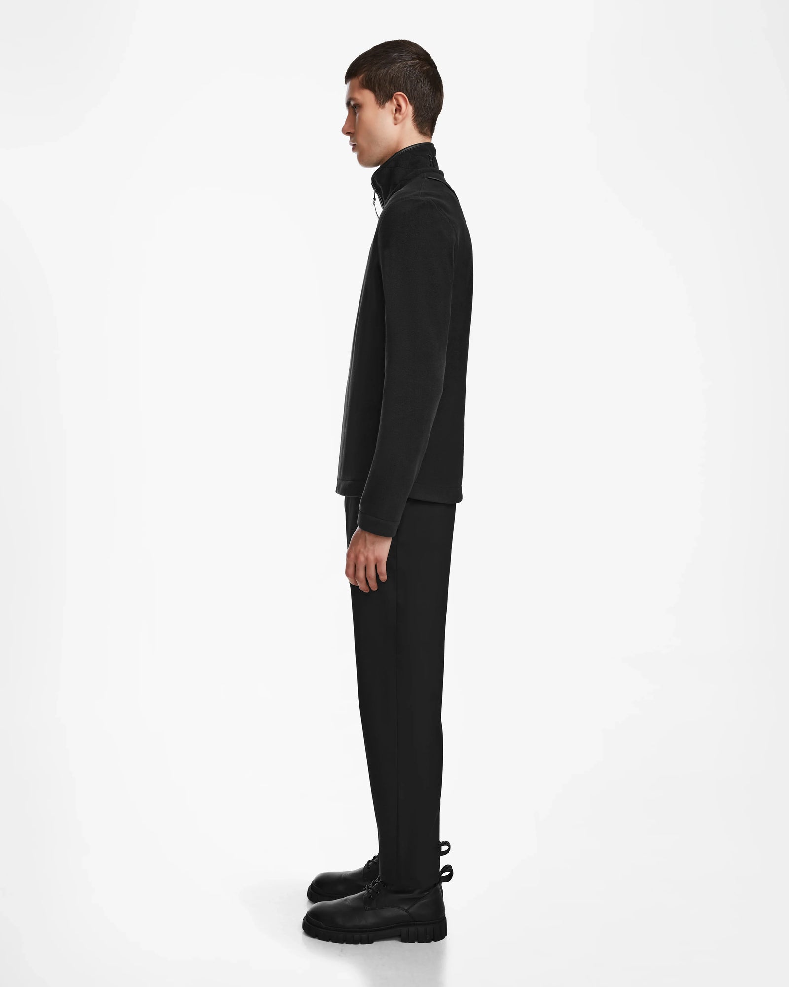 A young man stands in profile against a white background, wearing a black turtleneck, black pants, and the Rudsak ARAGON - 6524539 Black men’s fleece jacket with sleek vegan leather trims.