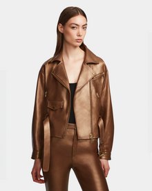 A woman with straight brown hair wears the Rudsak ARIELLE - 8124052 Antique Bronze lamb leather biker jacket and matching pants over a black top, standing against a plain white background.