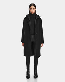 A woman with straight dark hair and a neutral expression models the Rudsak ARLA - 8124579 Black wool double-breasted coat over a black turtleneck, black faux leather pants, and ankle boots against a plain background.