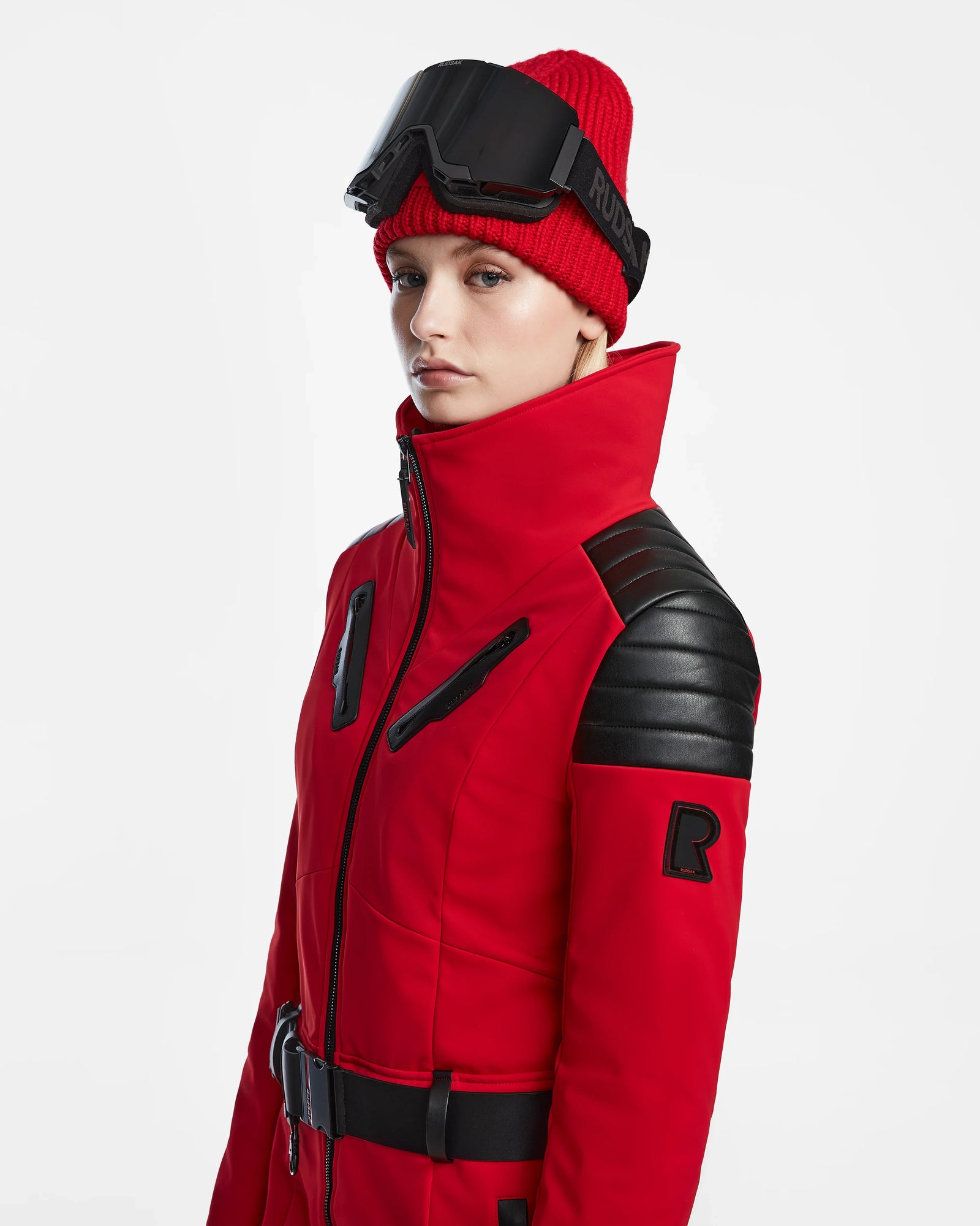 A person in the Rudsak ARLEY - 8124717 Flame Red ski jacket with black shoulder accents, a red knit beanie, and black ski goggles poses against a white backdrop, highlighting RECCO rescue technology.