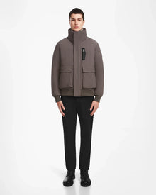 A man stands facing forward against a plain white background, wearing the Rudsak ASH - 6124599 Grey high-collar, water-resistant bomber jacket with large pockets, paired with black pants and black shoes.