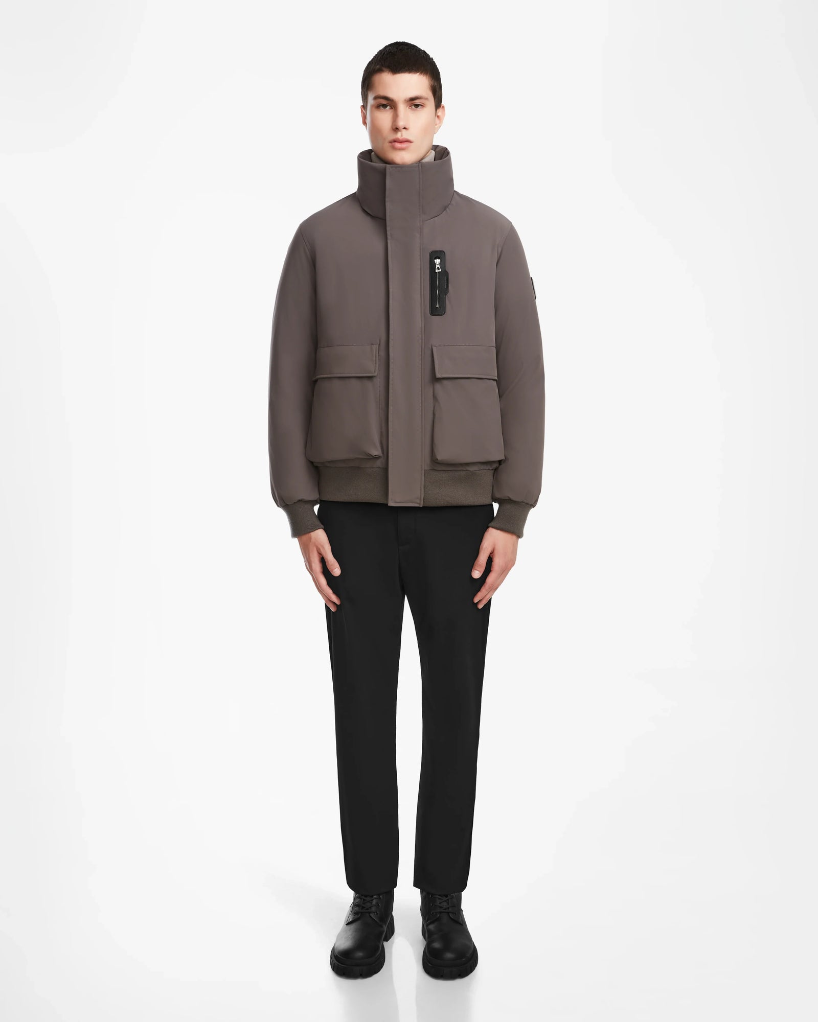 A man stands facing forward against a plain white background, wearing the Rudsak ASH - 6124599 Grey high-collar, water-resistant bomber jacket with large pockets, paired with black pants and black shoes.