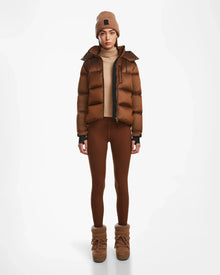 A woman wears the Rudsak ASHLYN S - 8124565 Antique Bronze women’s down puffer with RDS-certified down, a tan turtleneck, brown leggings and boots, plus a tan knit beanie, facing forward against a plain white background.