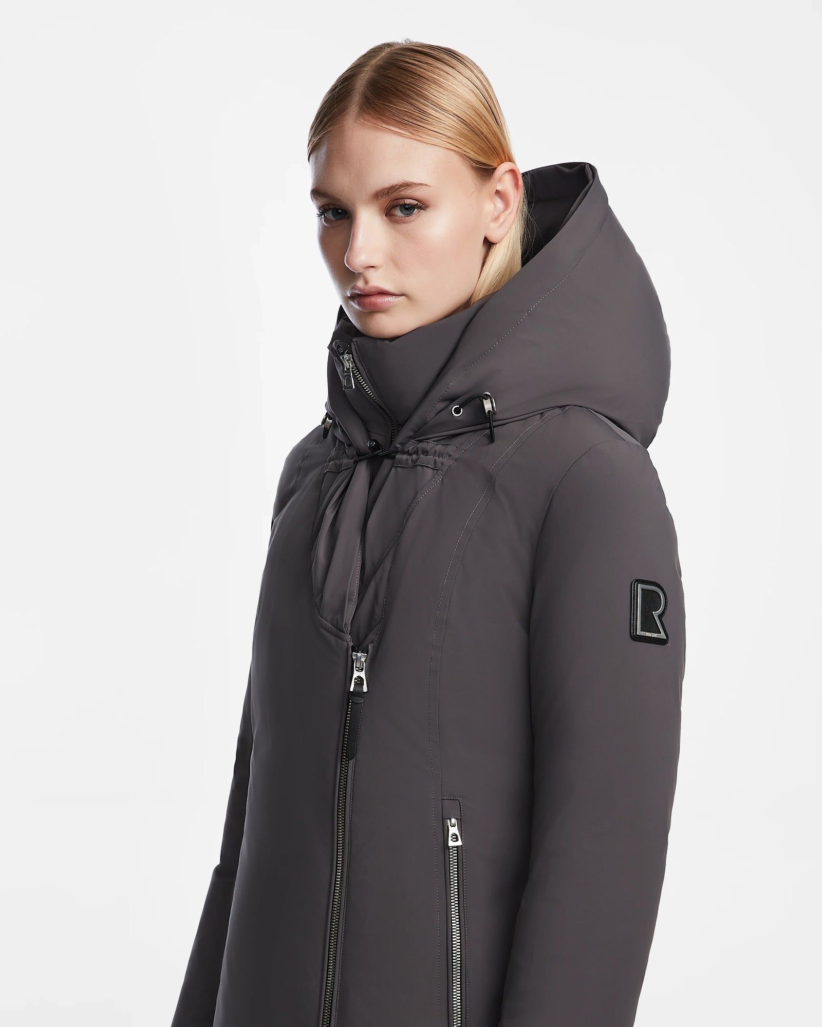 A woman with straight blonde hair models the Rudsak AURORE - L123561 Grey coat—a long, dark gray fitted winter style with a hood, high collar, double zipper, and an R patch on the upper sleeve—against a white background.