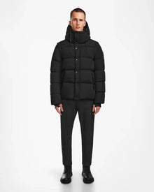 A man faces forward against a plain white background, wearing the Rudsak BOBBY - 6124506 Black mens down puffer jacket with a hood, paired with dark trousers and black shoes.