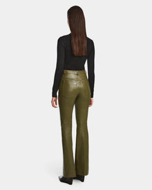 A woman with straight brown hair is seen from behind, wearing a black long-sleeve top and Rudsak CARISSA - 8524545 Loden Green high-waisted flare leather pants with black shoes, set against a white background.