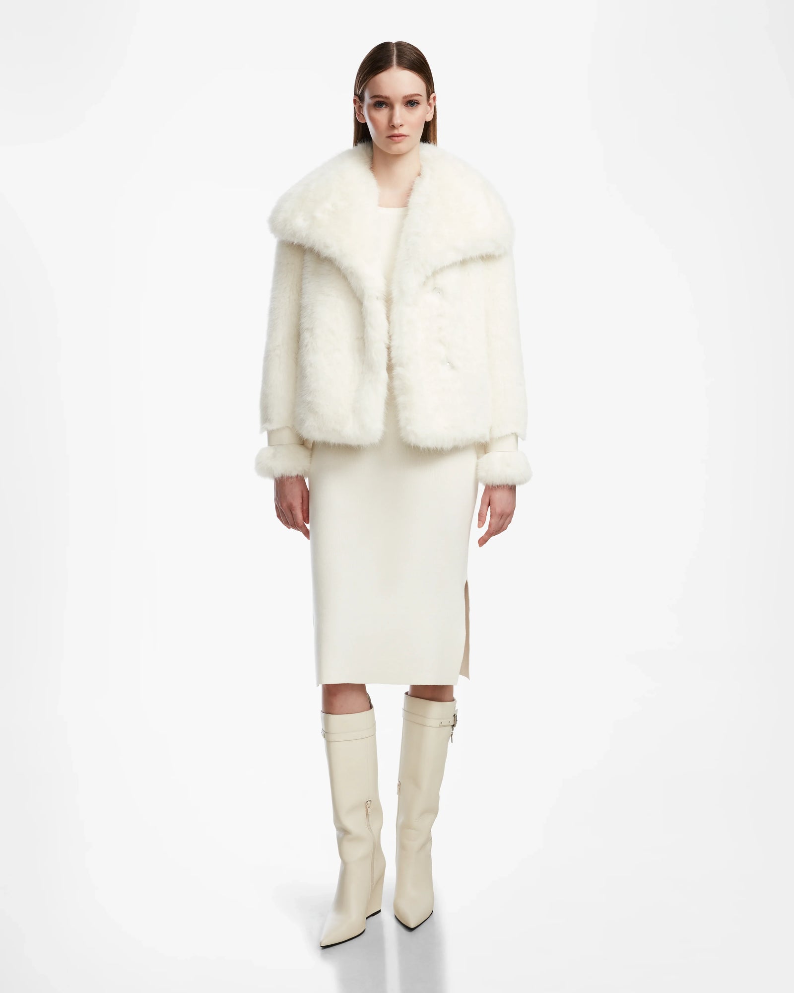 A woman with straight brown hair and a neutral expression stands against a white background, wearing the Rudsak CHLOE - L124564 Iceland faux fur jacket, a cream midi dress with side slit, and knee-high white heeled boots.