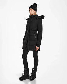 A person wearing the Rudsak CHRISTEL M-M - L124505 Black quilted winter coat with a fur-trimmed hood stands against a plain white background, styled with black leggings, black boots, and a black knit beanie.