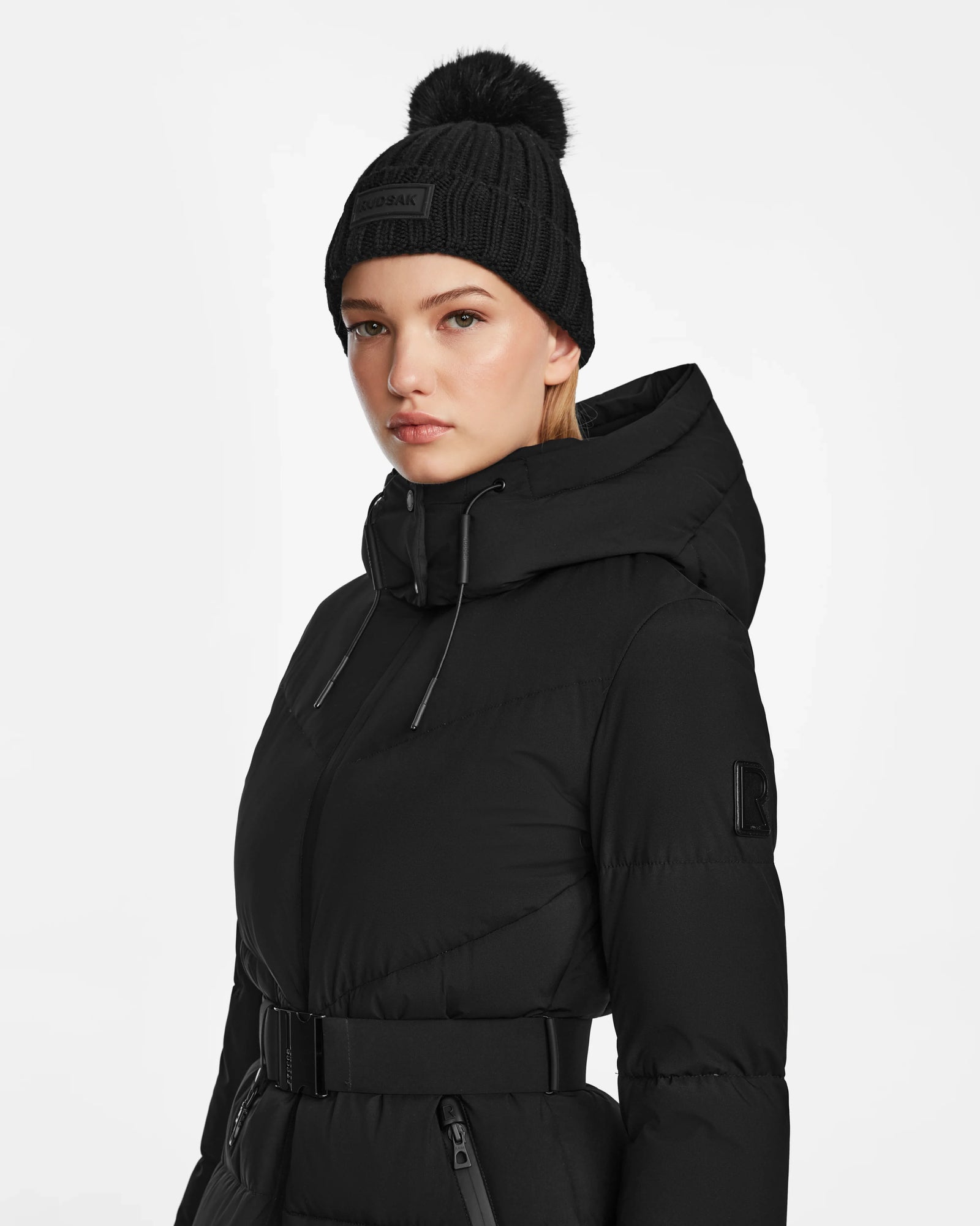 A woman wearing the Rudsak CHRISTEL M-M - L124505 Black quilted winter coat with a hood and a black pom-pom beanie, standing against a plain white background.