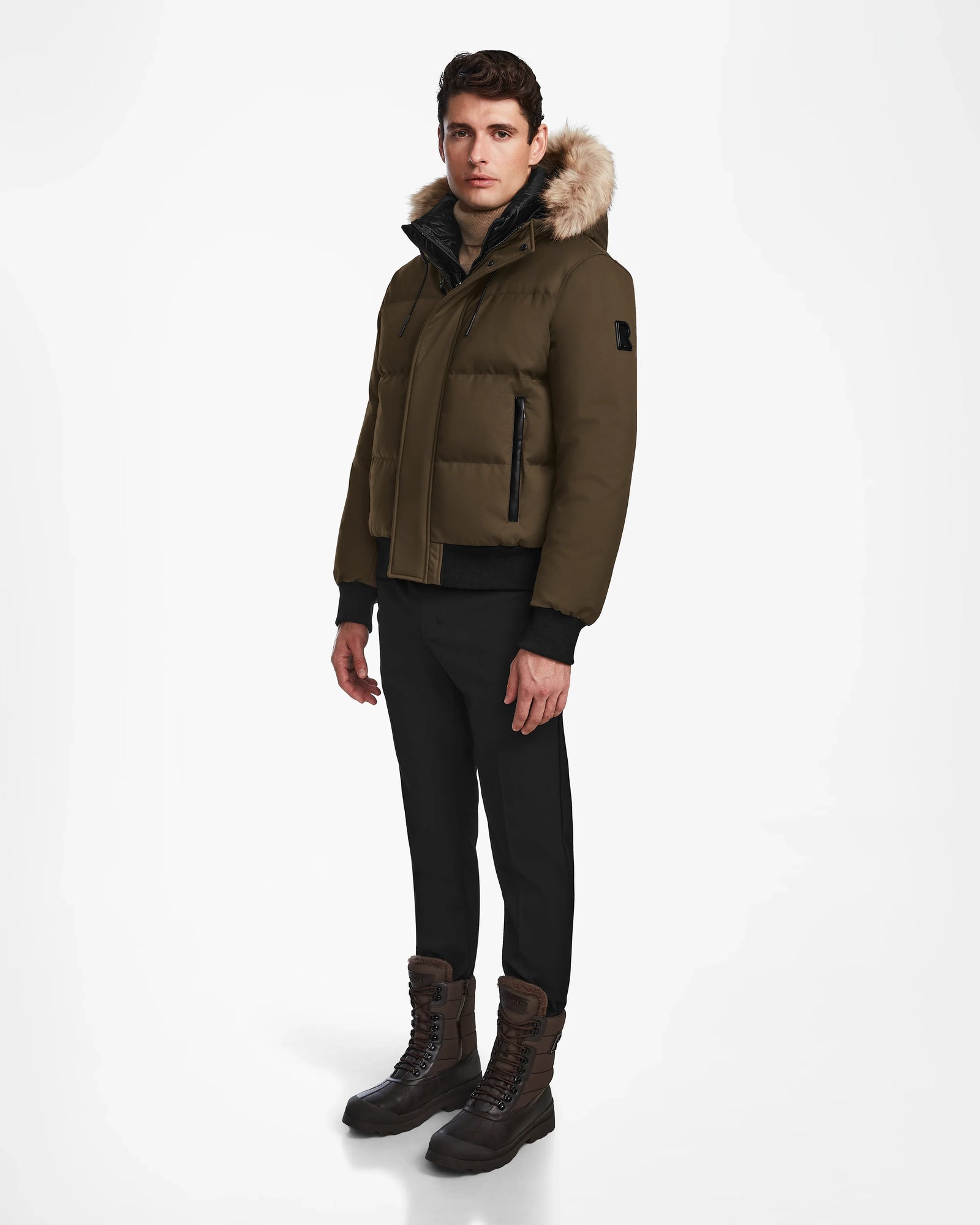 A man stands against a plain white background wearing the Rudsak DALLAS - M122501 Army weather-resistant puffer jacket with a fur-lined hood, paired with black pants and brown lace-up boots.