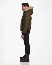 A man stands in profile against a white background, wearing the Rudsak DALLAS - M122501 Army weather-resistant puffer jacket with fur trim, black pants, and brown lace-up boots.