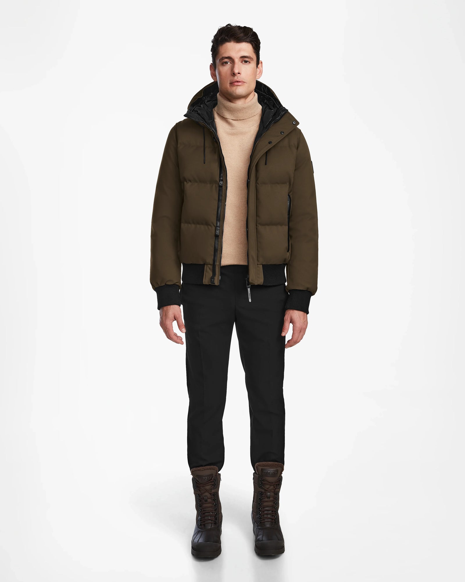 A man stands facing forward against a plain white background, wearing the Rudsak DALLAS - M122501 Army weather-resistant puffer jacket, a beige turtleneck sweater, black pants, and brown winter boots.