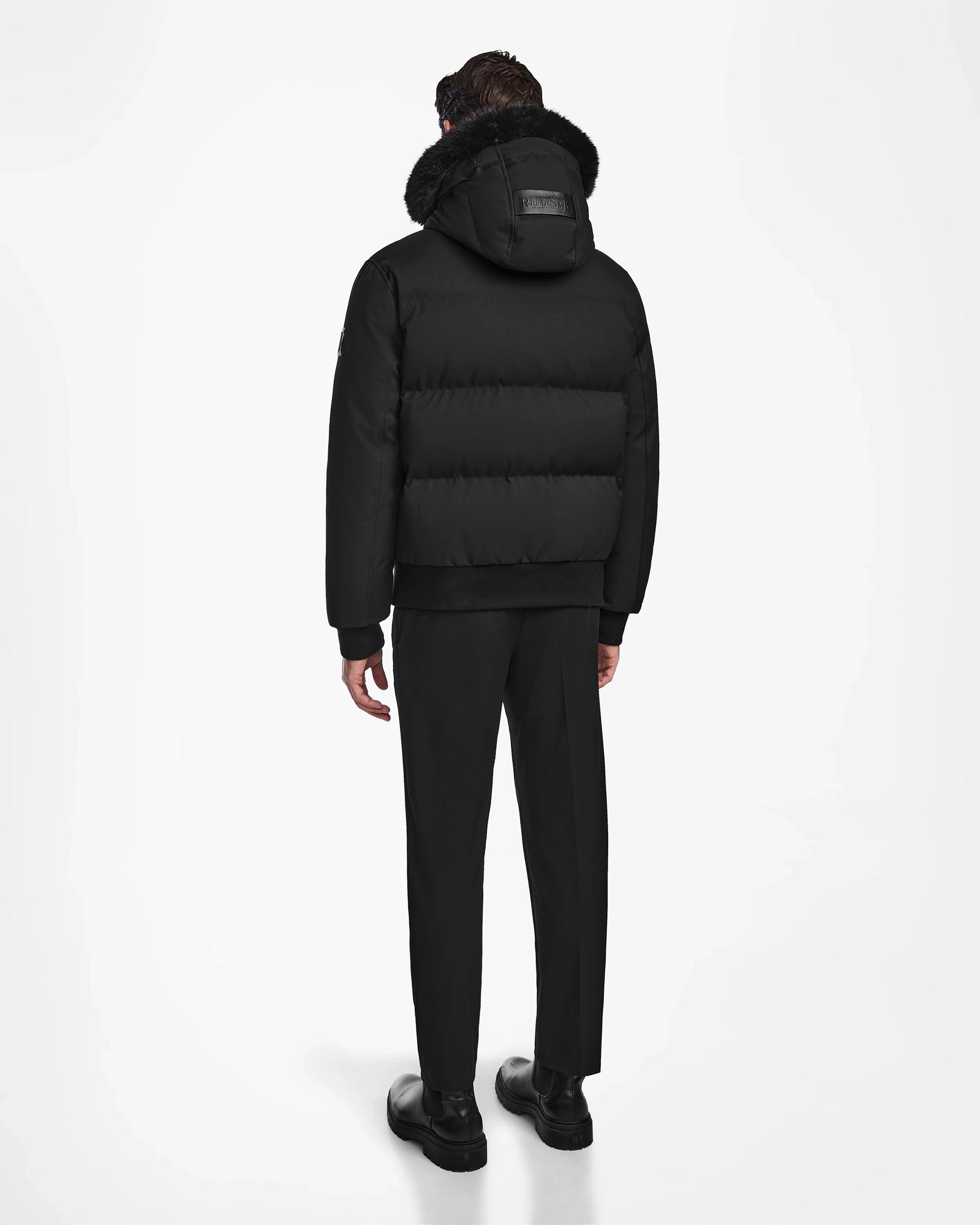 A person stands facing away, wearing the Rudsak DALLAS - M122501 Black weather-resistant puffer coat with fur trim, paired with black pants and shoes against a plain white background.
