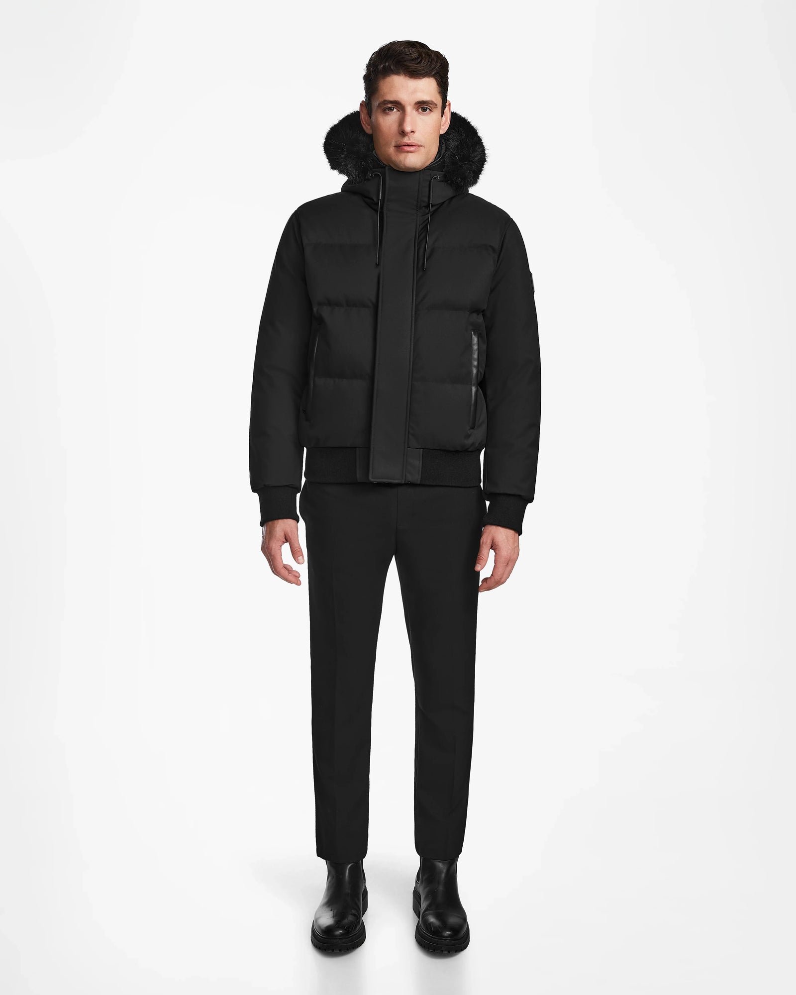 A man stands facing forward in a Rudsak DALLAS - M122501 black down bomber puffer coat with a fur-lined hood, paired with black pants and boots, set against a plain white background.