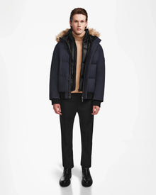 A person stands against a white background wearing the Rudsak DALLAS - M122501 Navy men's quilted down bomber with a fur-lined hood, paired with a beige turtleneck, black pants, and boots. The weather-resistant puffer offers warmth and style for winter.