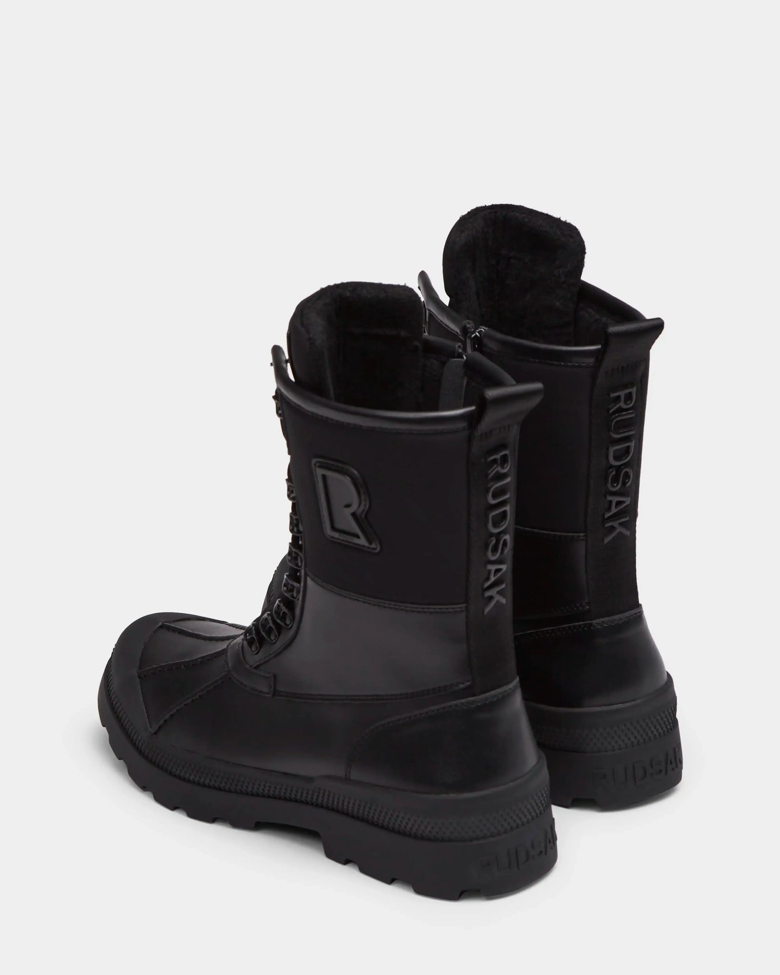 A pair of Rudsak DAMIEN - M222500 black men’s winter boots with insulated design, textured soles, side zippers, and bold RUDSAK branding on the back heel and sides, shown on a light gray background.