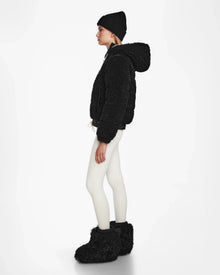 A woman stands in profile against a white background, wearing the Rudsak DANI S - 8124537 Black cropped ski jacket with a fuzzy hood, paired with a black beanie, white leggings, and large black fuzzy boots.