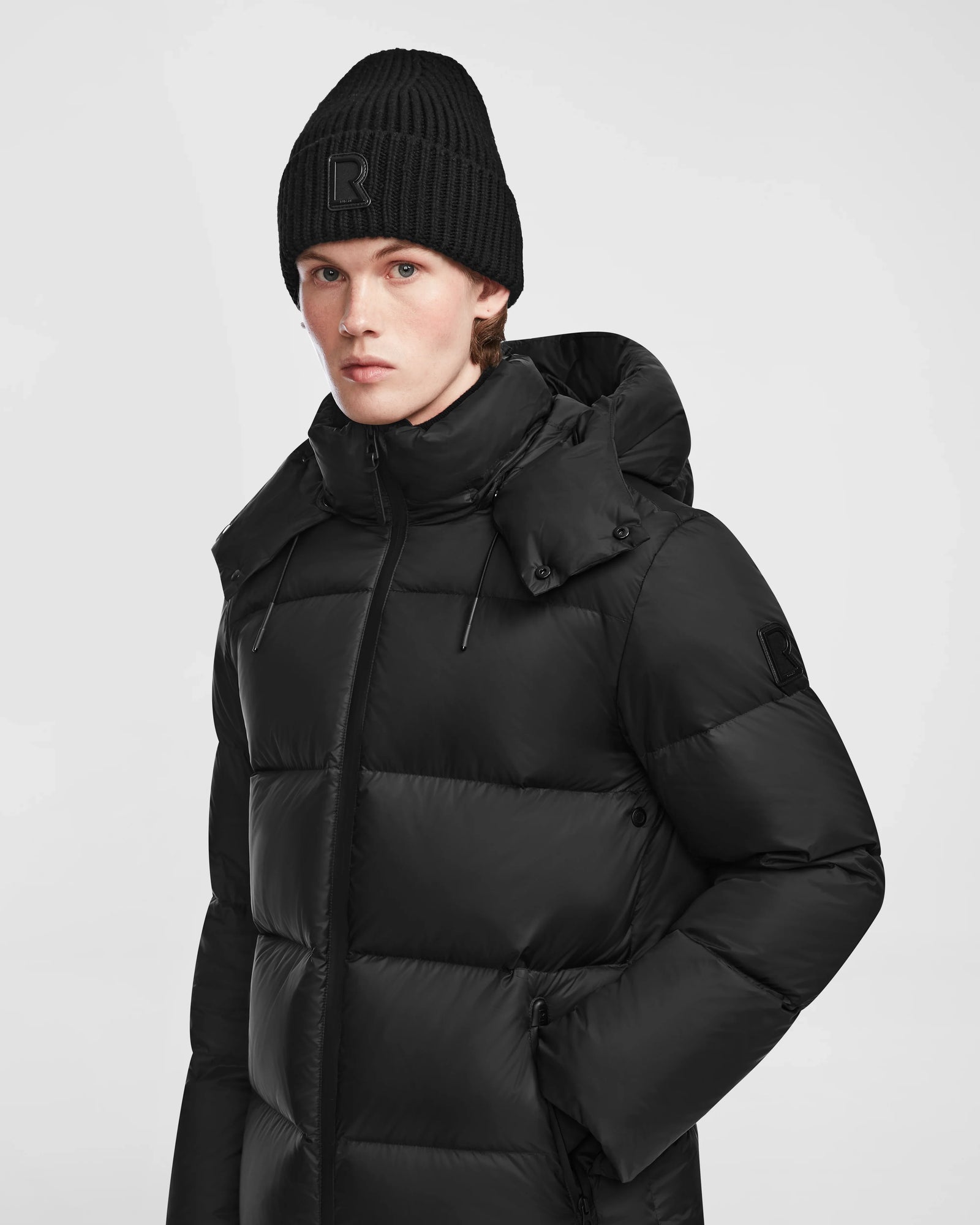 A person wearing the Rudsak DANIEL M - M124510 Black mens hooded down puffer and a black knit beanie stands against a plain light background, looking directly at the camera with a neutral expression.