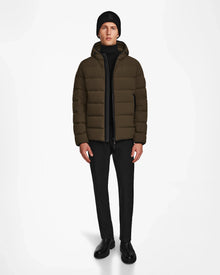A person stands against a plain white background wearing the Rudsak DEMIAN M - M123507 Army men’s down puffer jacket, made from sustainable recycled fabric, paired with black pants, boots, sweater, and a knit beanie.