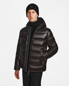 A man stands against a white background wearing the Rudsak DEMIAN S - M123508 Black down puffer jacket, paired with a black knit beanie and black pants, with one hand in his pocket.