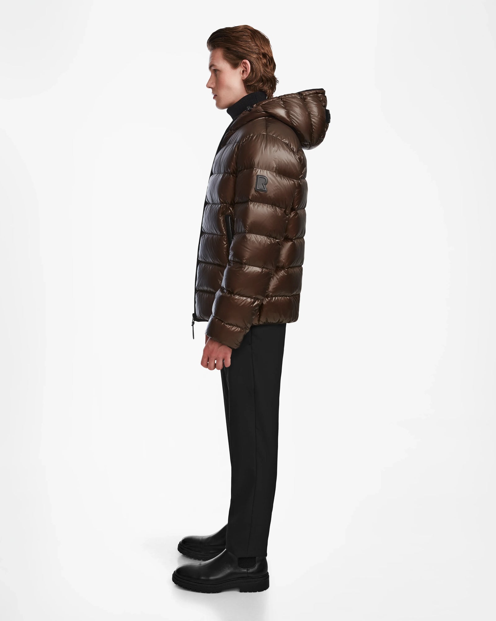 A man stands in profile against a white background, wearing the Rudsak DEMIAN S - M123508 Espresso mens down puffer jacket, paired with black pants and chunky black shoes.