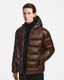 A young person models the Rudsak DEMIAN S - M123508 Espresso men’s down puffer jacket, with premium down fill, black turtleneck, and pants, standing against a plain white background and looking directly at the camera.