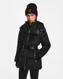 A woman wearing the Rudsak DONIA - L124563 Black convertible winter coat, a black knit hat with an R, black gloves, and black leggings stands against a plain white background.