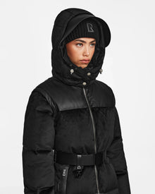 Wearing the Rudsak DONIA - L124563 black convertible winter coat with leather accents and metallic buttons, plus a black R-marked beanie and wide belt, they face slightly left against a white background looking serious.