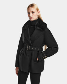 A woman with long blonde hair models the Rudsak ELENOR - 8124707 Black trench coat with a shearling collar, styled with a black turtleneck and pants, against a plain light gray background.