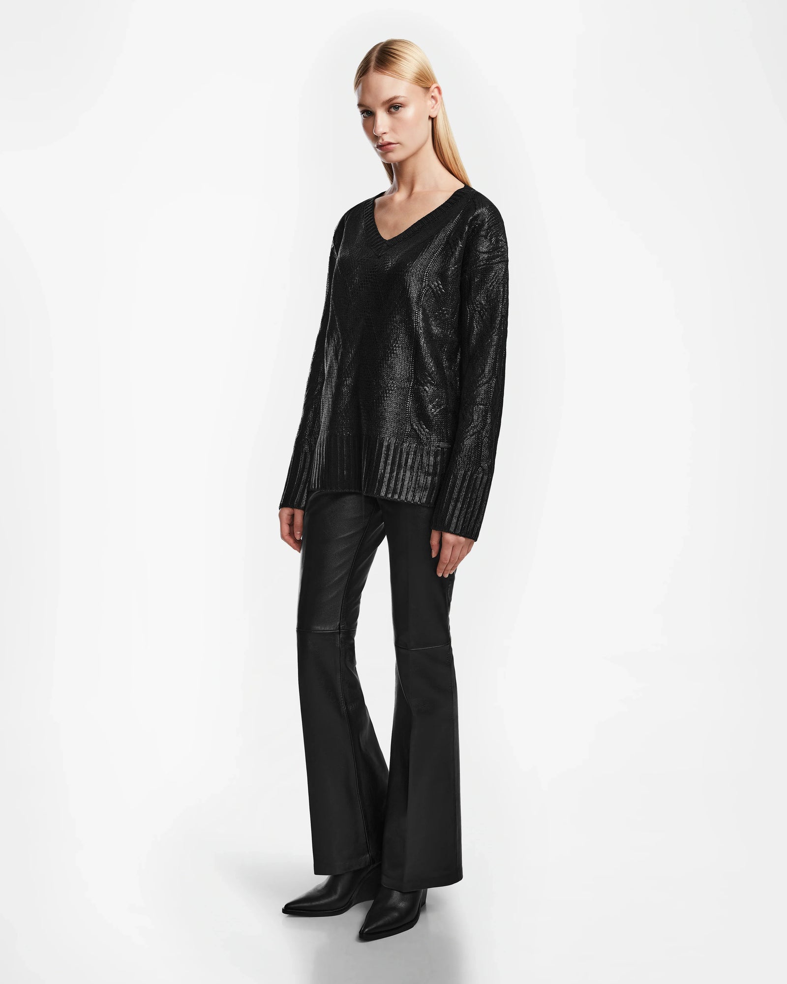 A woman with straight blonde hair wears the Rudsak ELLE - 8524597 Black V-neck sweater, black flared pants, and pointed black boots, standing against a plain white background.